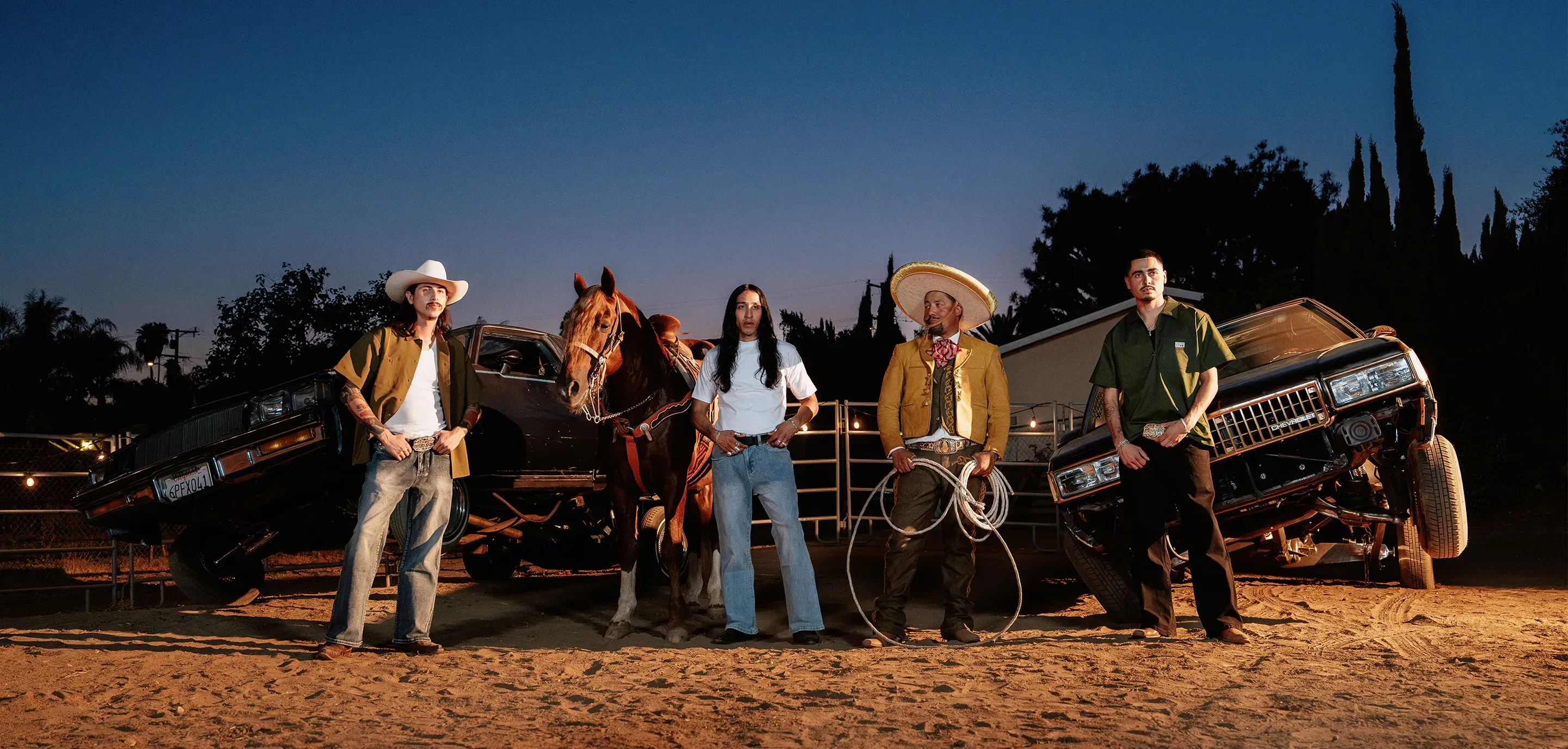 Four men in western wear, a charro suit and white tees pose at dusk beside a horse and two lowriders under string lights.