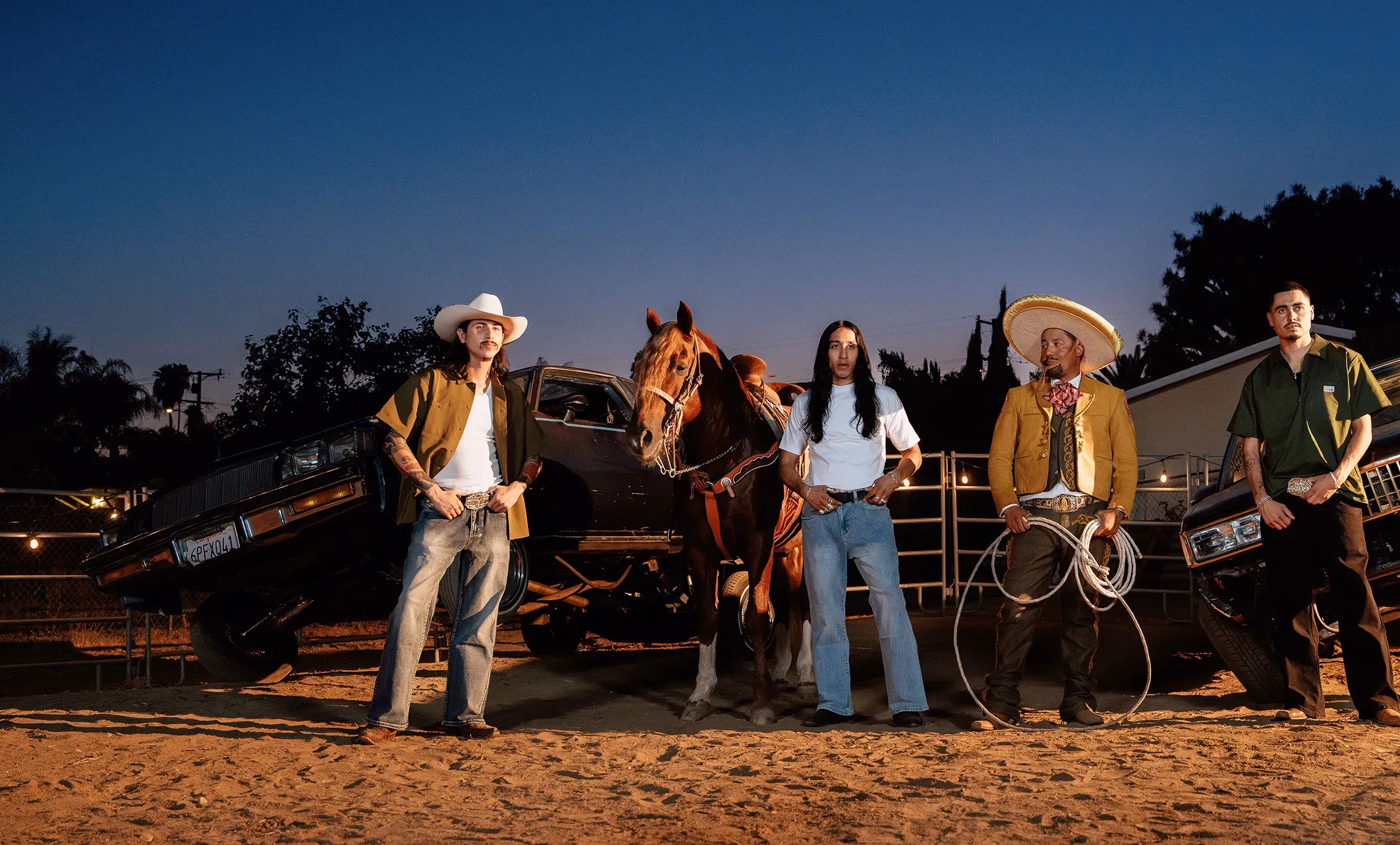 Four men in western wear, a charro suit and white tees pose at dusk beside a horse and two lowriders under string lights.