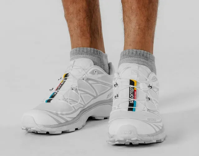 Close-up of two feet in grey ankle socks and white running sneakers against a light grey backdrop.