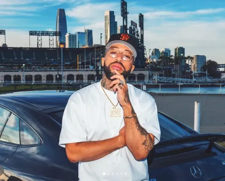 Larry June in a white t-shirt and SF cap leaning on a car