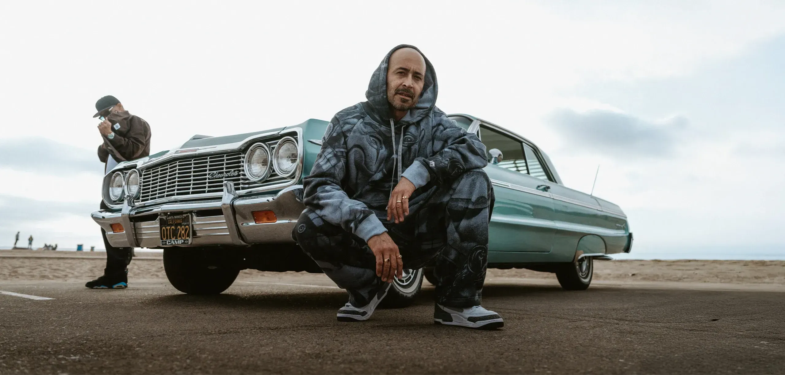 Mister Cartoon crouched beside a teal lowrider — Pro Club limited-edition graphics collab