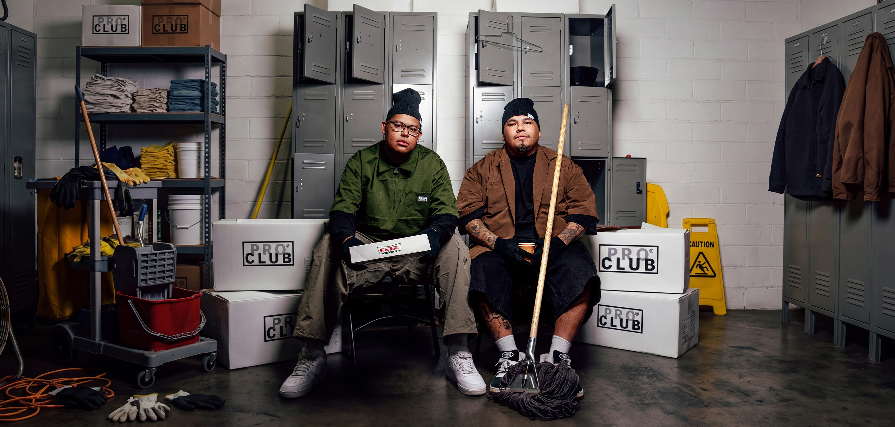 Two men in Pro Club workwear and black beanies sitting in a janitorial locker room, one in an olive green shirt holding a Krispy Kreme box and one in a brown shirt holding a mop, surrounded by Pro Club boxes and cleaning supplies