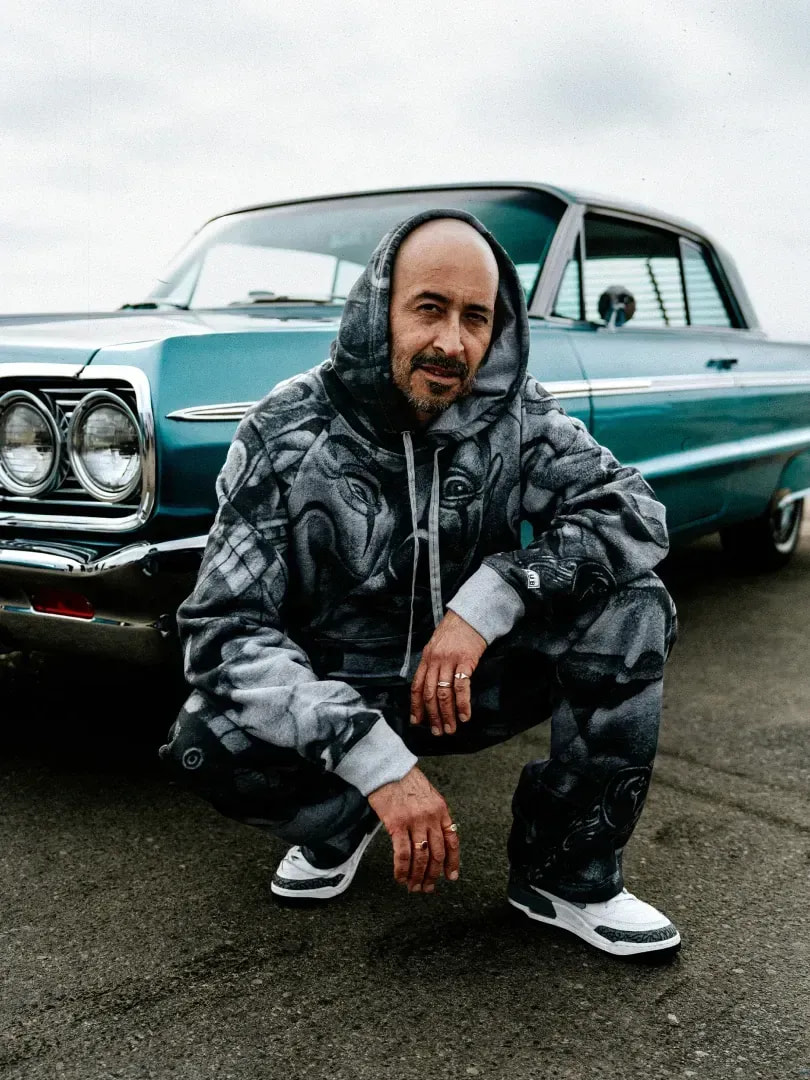 Man in an airbrushed grey Pro Club hooded tracksuit by a teal lowrider