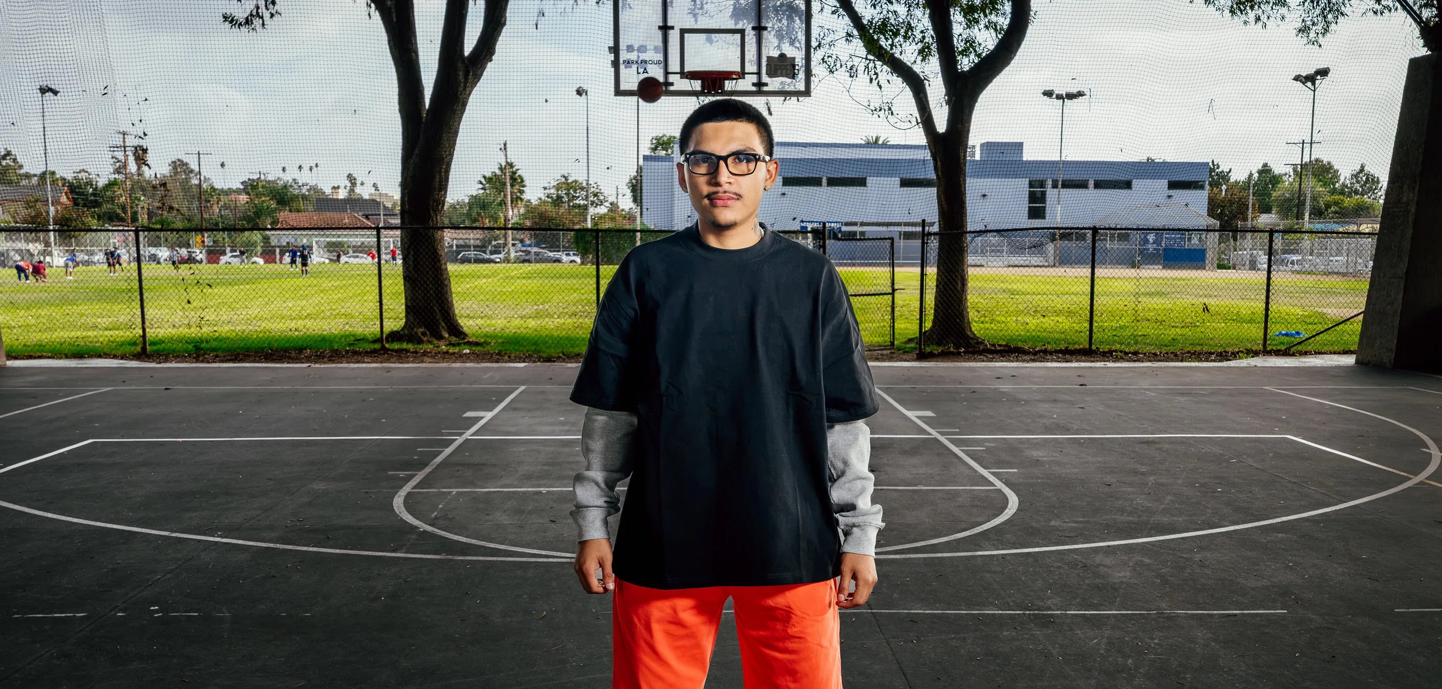 ProClub man in black oversized t-shirt with grey thermal layered sleeves and orange sweatpants standing on an outdoor basketball court