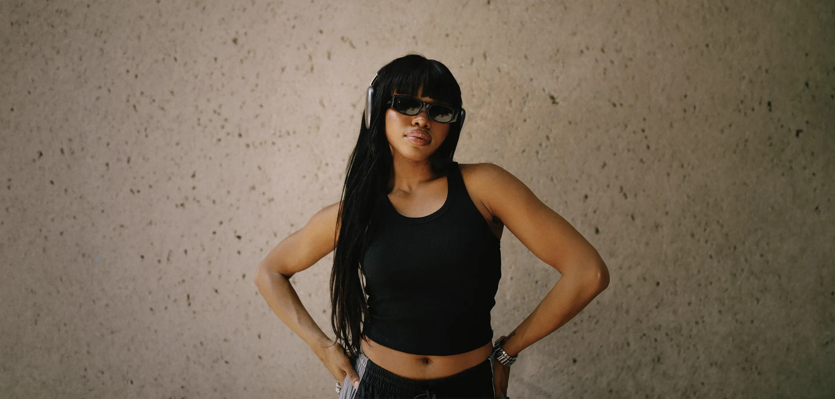Woman in a Pro Club black ribbed tank top and gray drawstring pants wearing black sunglasses, silver watch, and over-ear headphones, posing with hands on hips against a textured concrete wall
