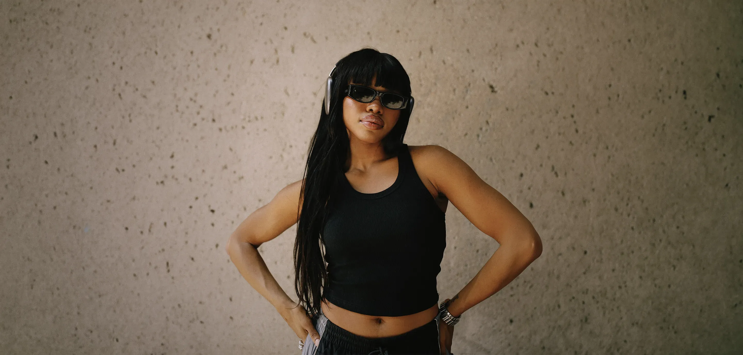 Woman in a Pro Club black ribbed tank top and gray drawstring pants wearing black sunglasses, silver watch, and over-ear headphones, posing with hands on hips against a textured concrete wall