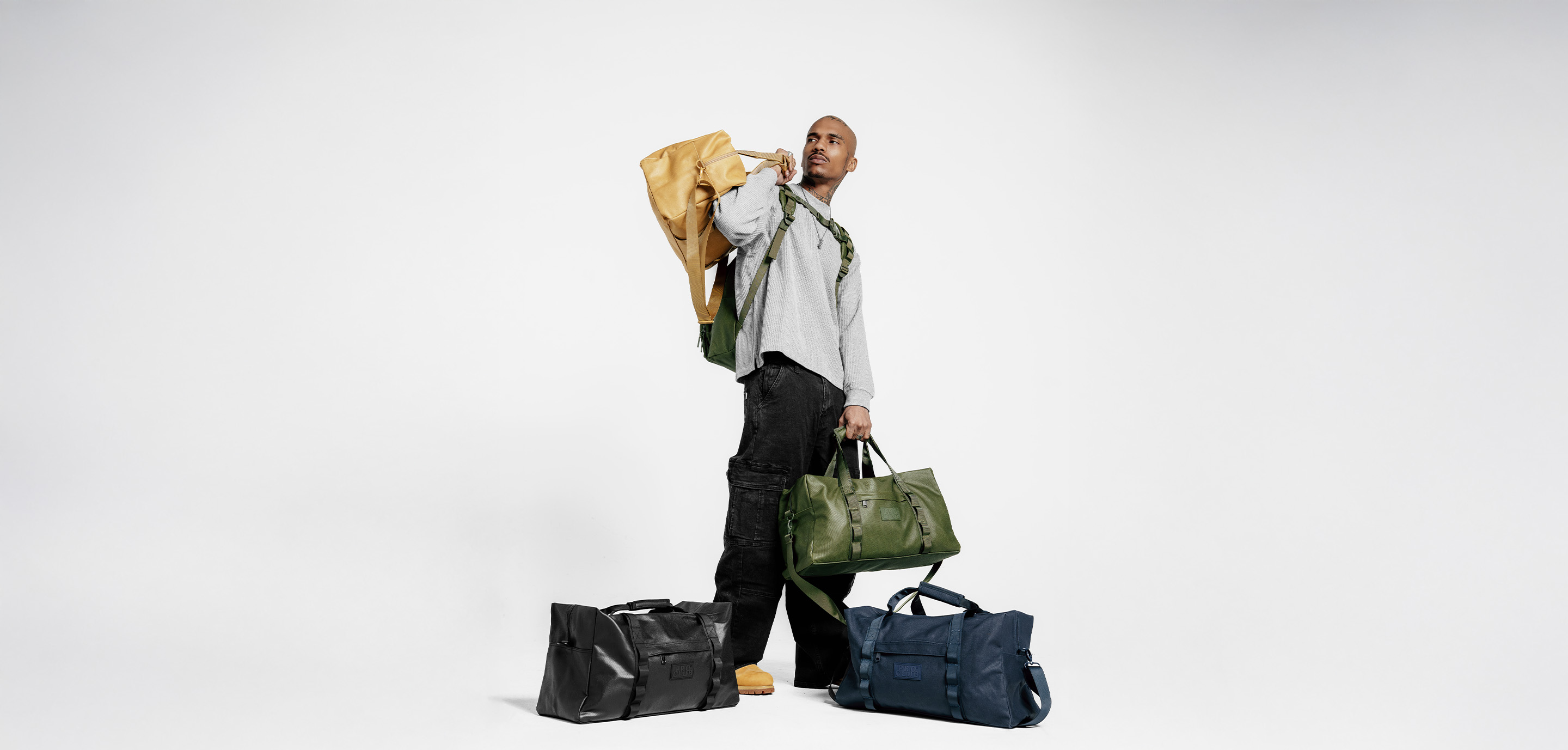 Man in a gray waffle-knit sweater and black cargo pants carrying a tan and olive green Pro Club backpack over his shoulder and an olive duffle bag, with black and navy Pro Club duffle bags on the floor against a white studio background