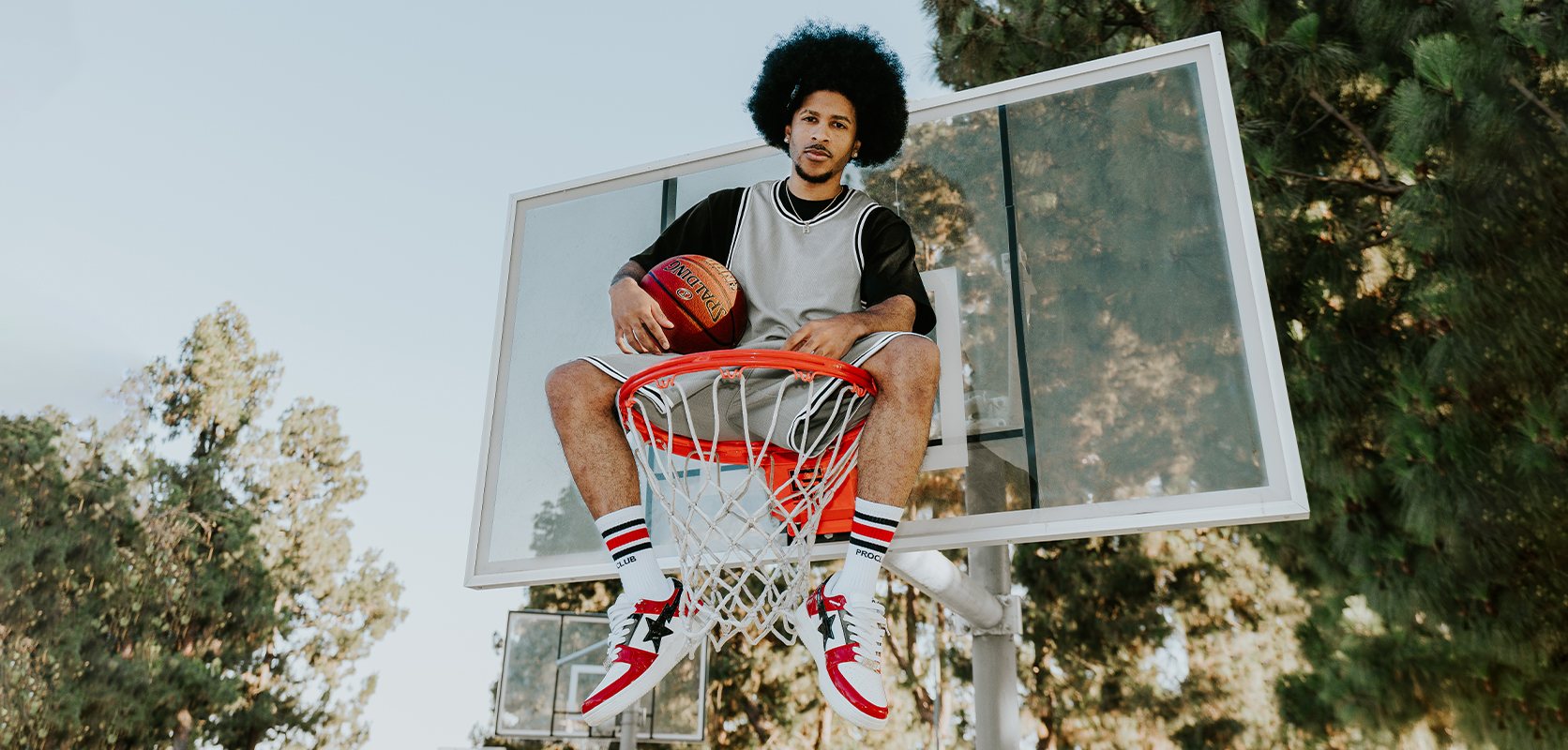 Man in a grey Pro Club basketball jersey sitting on a basketball rim holding a ball