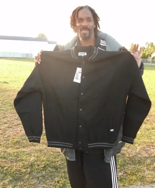 Snoop Dogg holding up a black Pro Club varsity jacket in a field