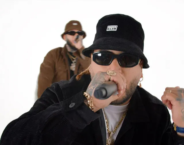A man in a black corduroy bucket hat with a Pro Club patch and dark sunglasses speaks into a microphone, with a second man in a brown bucket hat behind him.