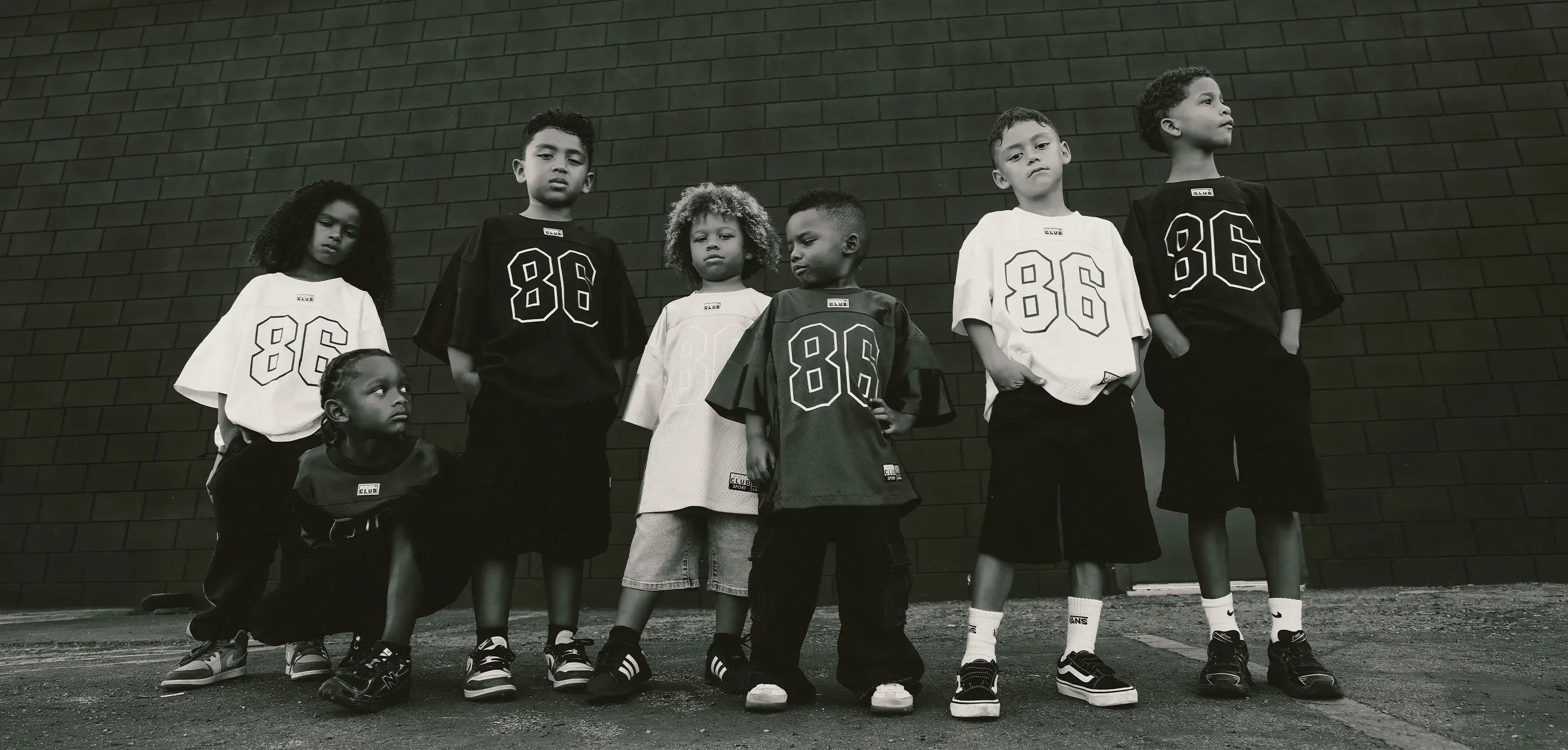 Group of seven kids wearing Pro Club "86" football jerseys in black and white, posing together against a dark brick wall in a black-and-white photo