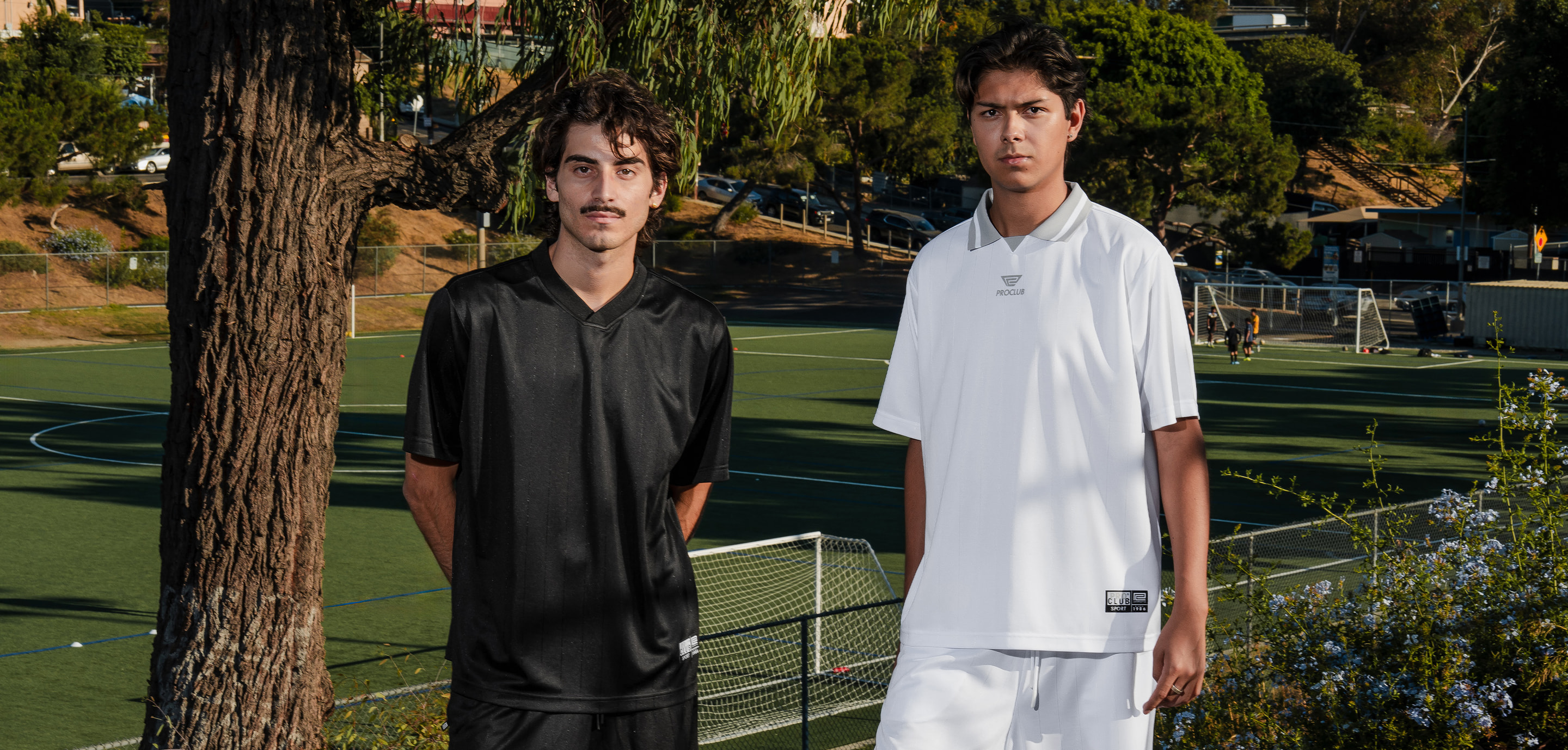 Two men in black and white Pro Club soccer jerseys beside a turf soccer field
