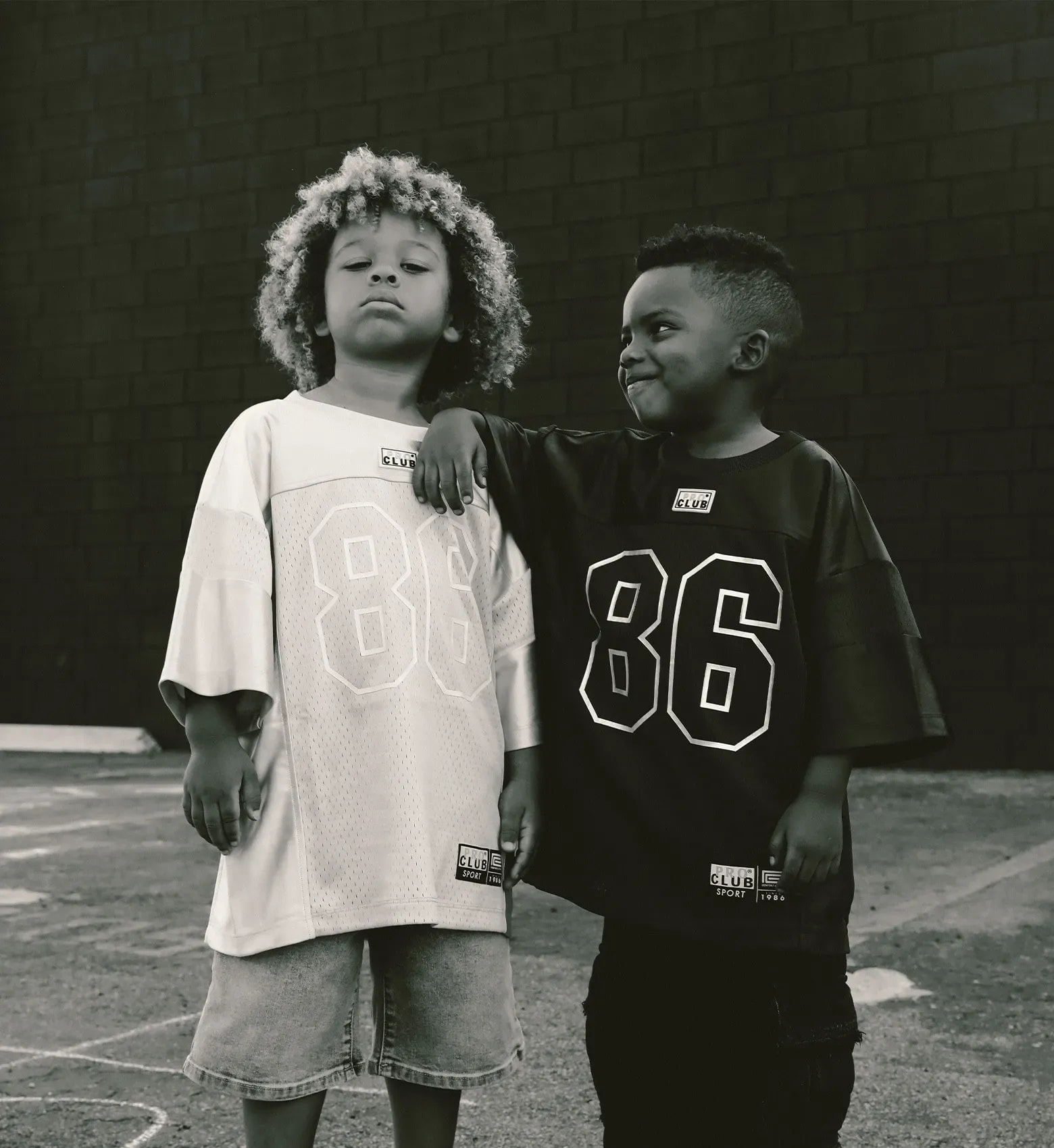 Two young boys in black and grey velour track sets and gold chains crouch against a white wall, each holding a handful of cash.