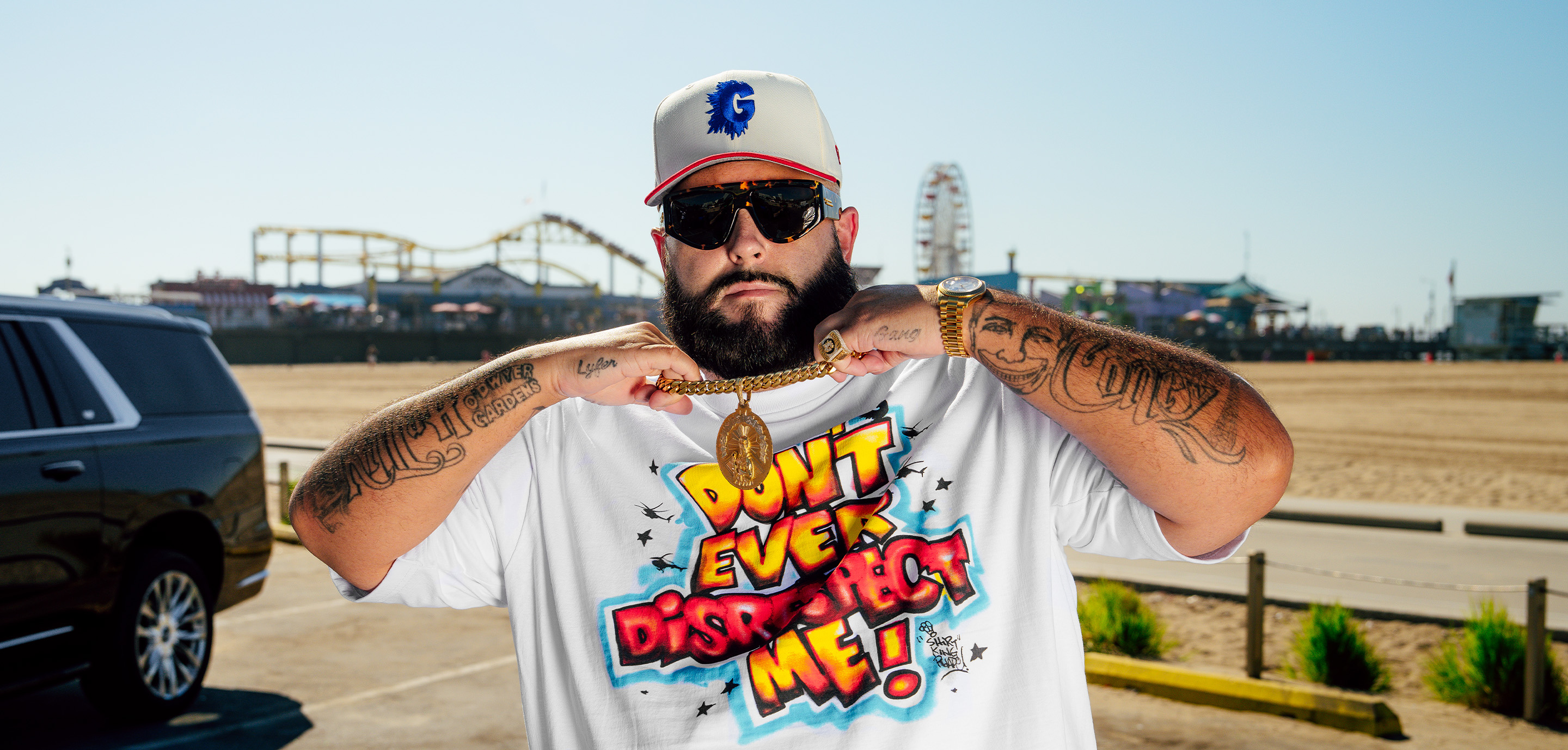 ProClub man wearing a white graphic t-shirt with ‘Don’t Ever Disrespect Me’ airbrush-style print, gold chain, cap, and sunglasses at Santa Monica Pier